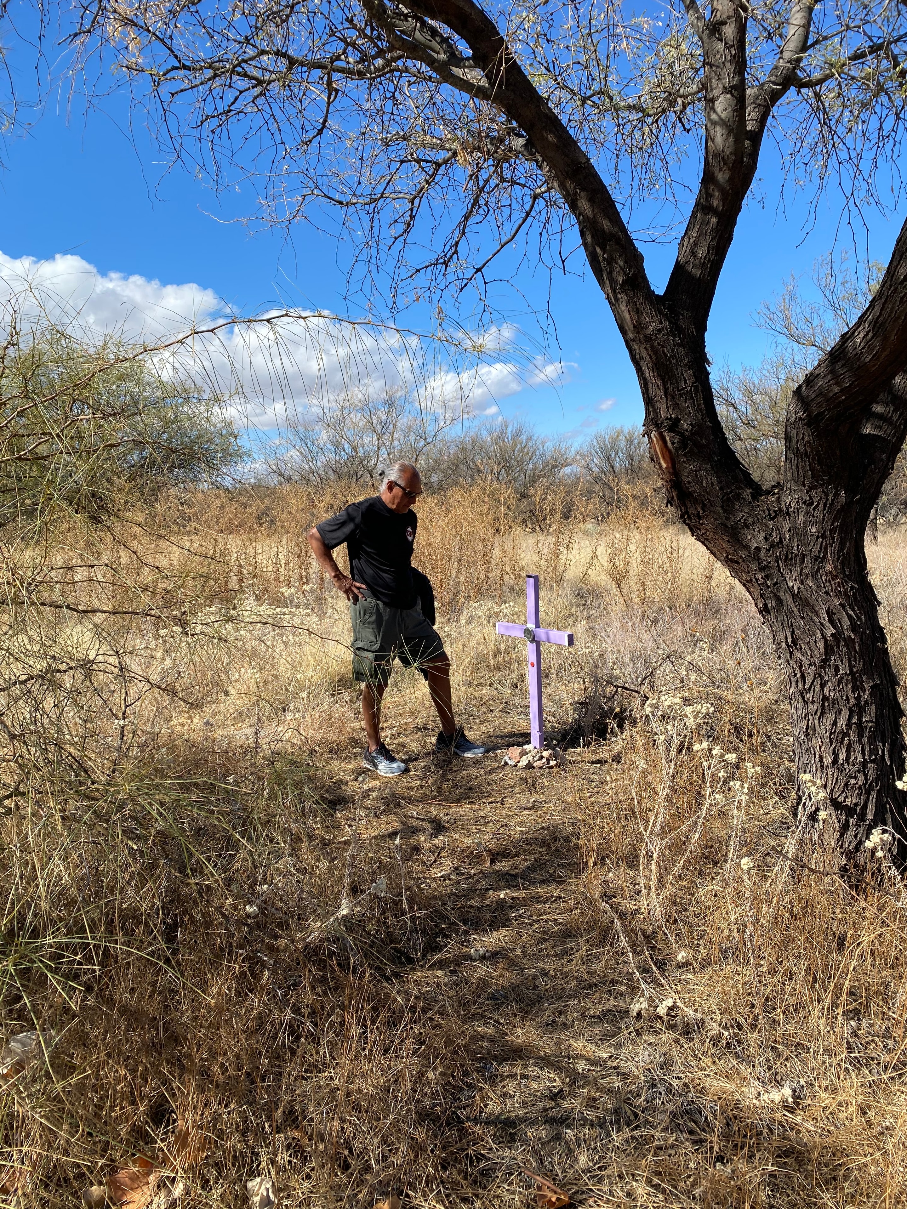 Kino Springs — Ricardo Santos Hernández stands beside a hand-crafted lavender cross placed by Alvaro Enciso's team of Samaritans in the desert scrub of Kino Springs, southern Arizona — the exact location, near Ricardo's home, where a migrant perished. A mesquite tree arches overhead, the dry grass and open sky bearing witness. Each cross is placed with precision, guided by navigational data that leads the team to the spot where a human being took their last breath. A small, handmade marker in an immense and indifferent landscape. Art as witness. Humanity as resistance. by Ricardo Santos Hernández