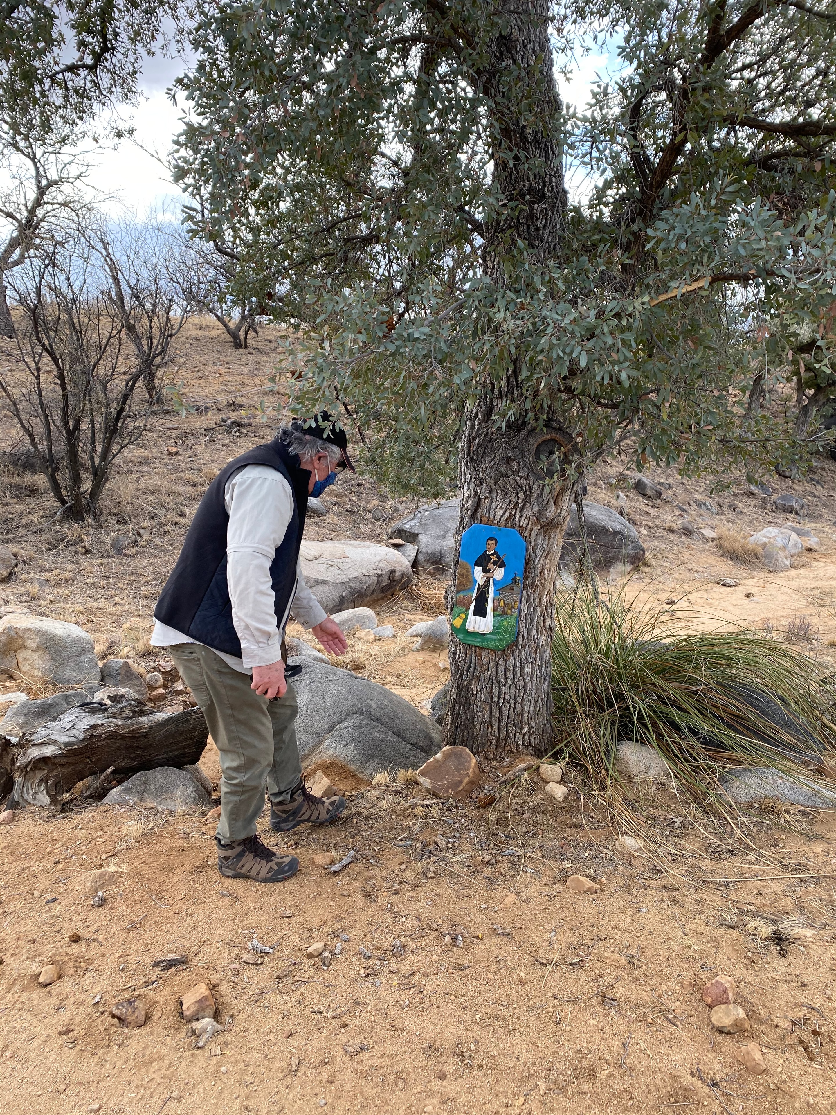 San Martín in the Patagonia Mountains — Alvaro Enciso pauses before a retablo of San Martín de Porres placed by an anonymous hand on a live oak tree along a trail in the foothills of the Patagonia Mountains in southern Arizona, near the US-Mexico border. The hand-painted folk devotional — a quiet act of faith left in the desert — speaks to the same humanity that drives Alvaro's cross-placing work: the belief that every life that has passed through this landscape deserves to be seen and honored. by Ricardo Santos Hernández