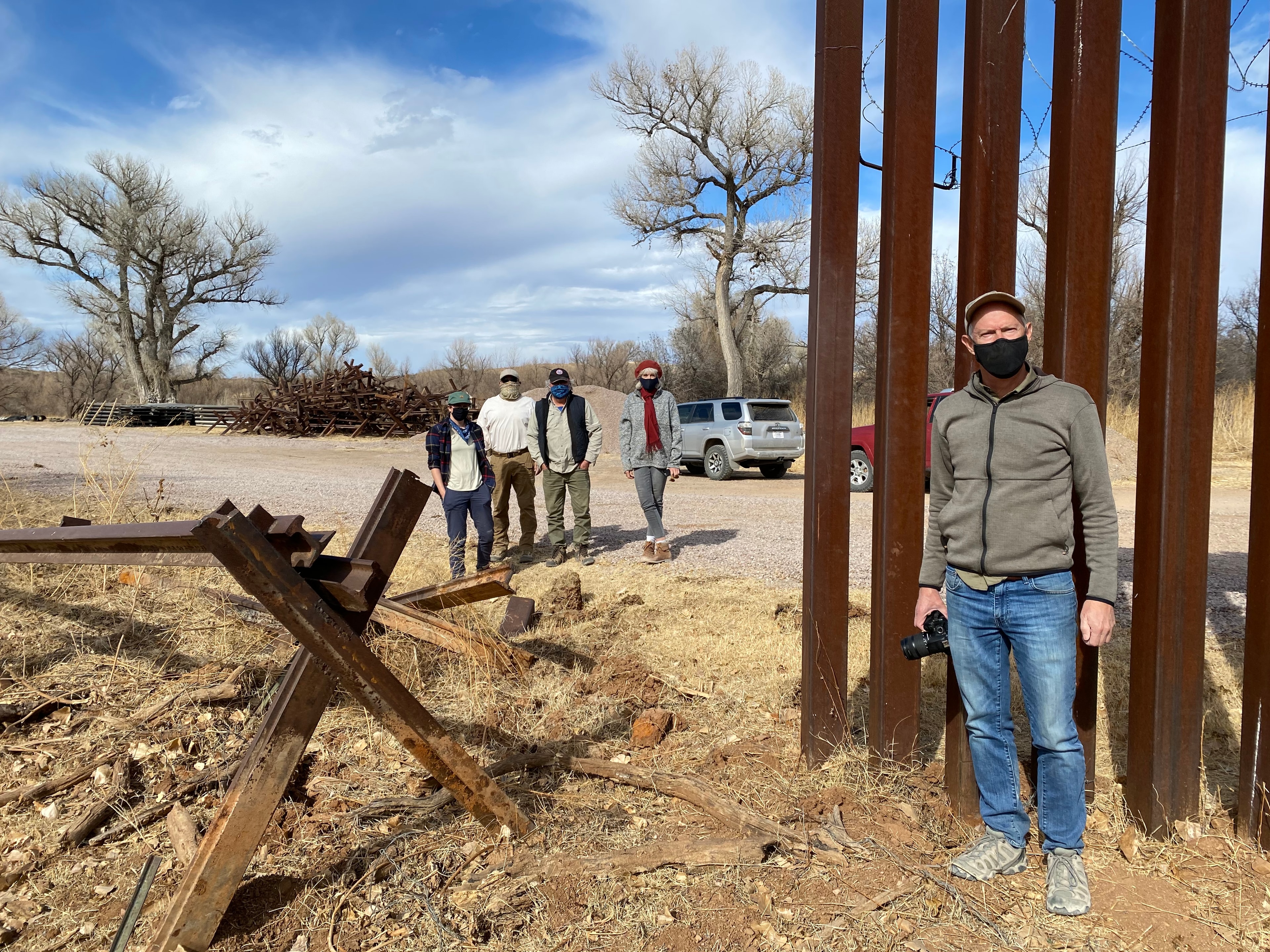 The Cross Man at the Santa Cruz River — Alvaro Enciso, known as the Cross Man, and his team at the Santa Cruz River's point of entry into Arizona along the US-Mexico border. Using an advanced navigational system, Alvaro guides his team to the exact locations where migrants have perished in the desert, placing hand-crafted wooden crosses to honor each life lost. The rusted steel bollards and tank trap barriers of the border wall frame the landscape behind them — a monument to policy set against a monument to humanity. Ricardo Santos Hernández has collaborated with Alvaro on this project and has included his artwork in exhibitions curated in southern Arizona. by Ricardo Santos Hernández