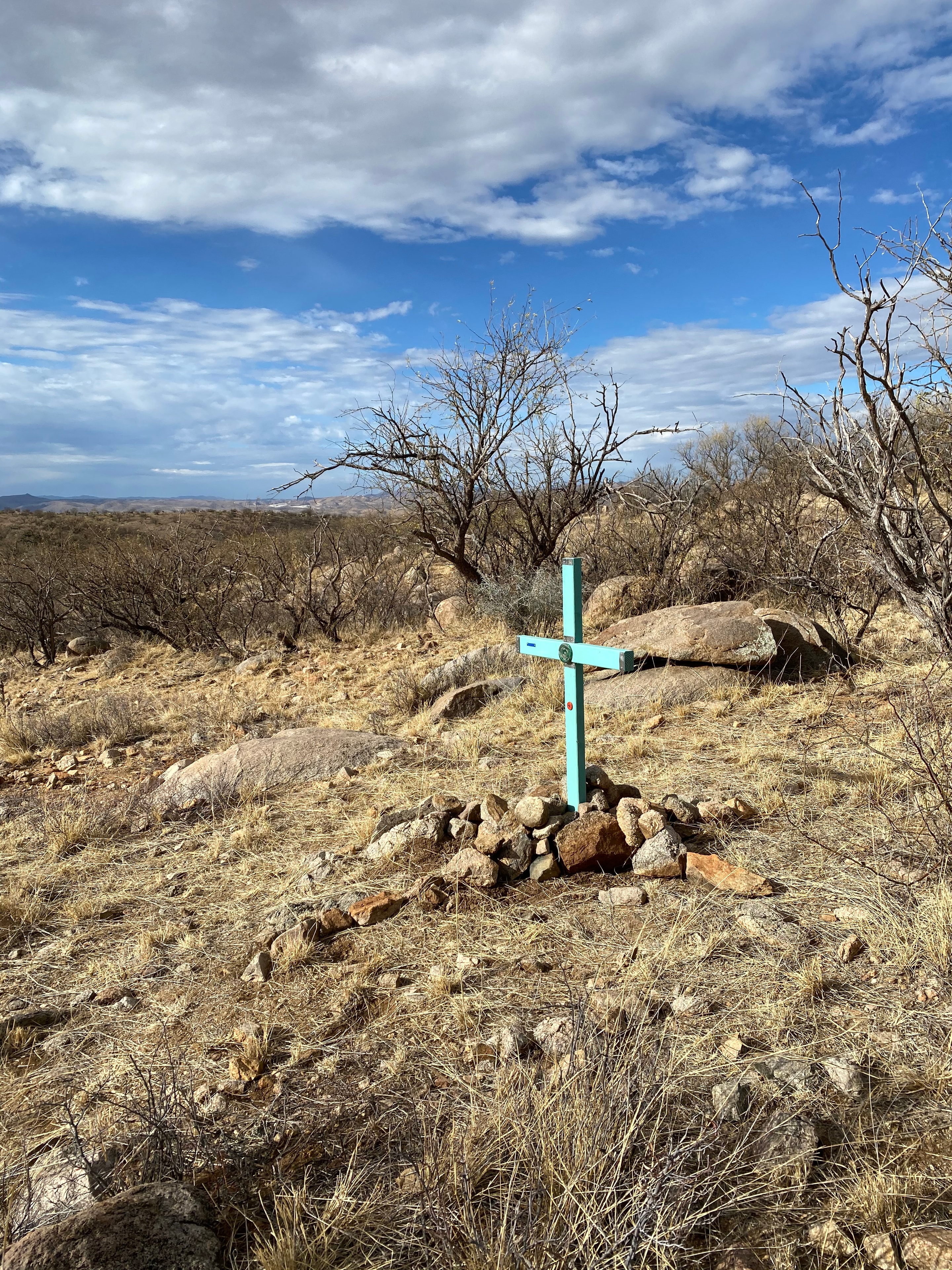 Patagonia Mountains — A solitary turquoise cross stands in the rocky hillside of the Sonoran desert in the foothills of the Patagonia Mountains, near the US-Mexico border. Placed by Alvaro Enciso's team of Samaritans and Ricardo Santos Hernández to honor a migrant who perished at this exact location. The vast desert sky opens behind it — bare mesquite, dry grass, ancient stone — the immensity of the landscape making the small handmade cross all the more profound. One life. One marker. The desert as witness. by Ricardo Santos Hernández