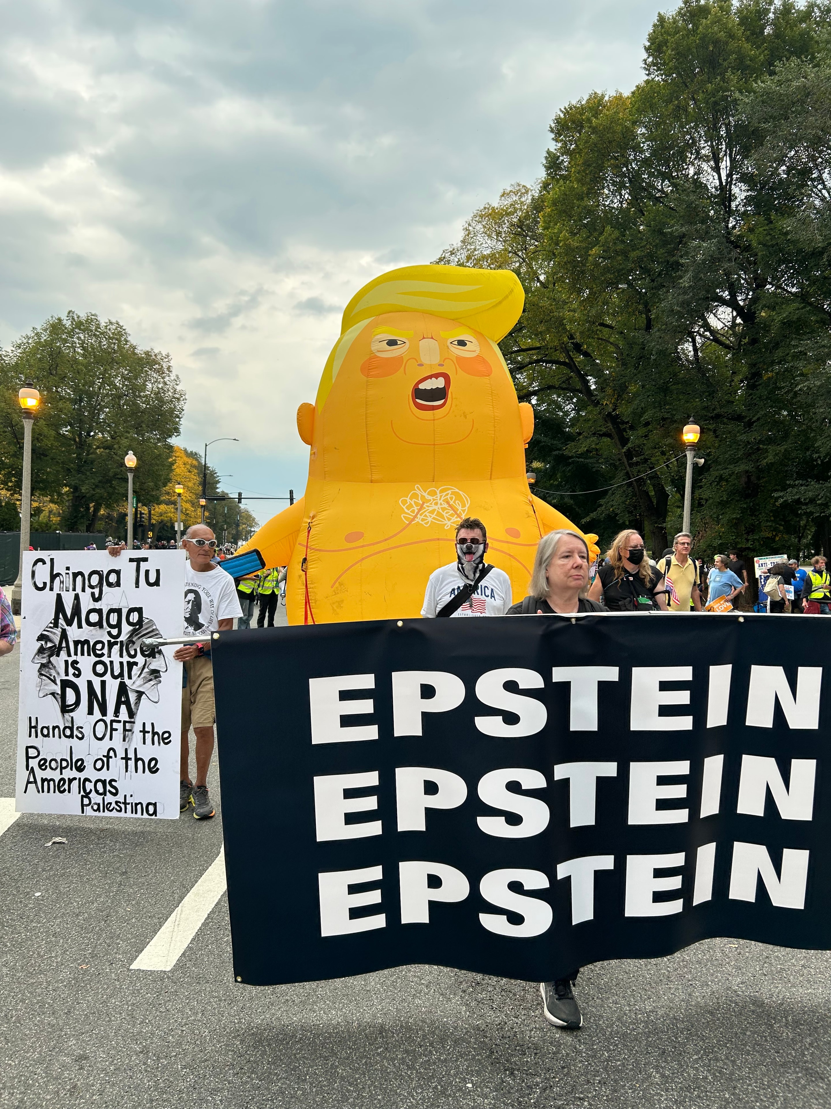 Epstein — Ricardo Santos Hernández, Chicago, September 2025. A documentary photograph from the No Kings March through the streets of downtown Chicago. Ricardo, a woman carrying a bold black-and-white EPSTEIN banner, and a group of Chicago lawyers pulling a giant inflatable Trump balloon walk hand in hand through the streets — Ricardo's sign reading "Chinga Tu MAGA / América is our DNA / Hands OFF the People of the Américas / Palestina" visible in the foreground. Artists, lawyers, and citizens united in the streets, demanding accountability. Art as witness. Art as demand. by Ricardo Santos Hernández