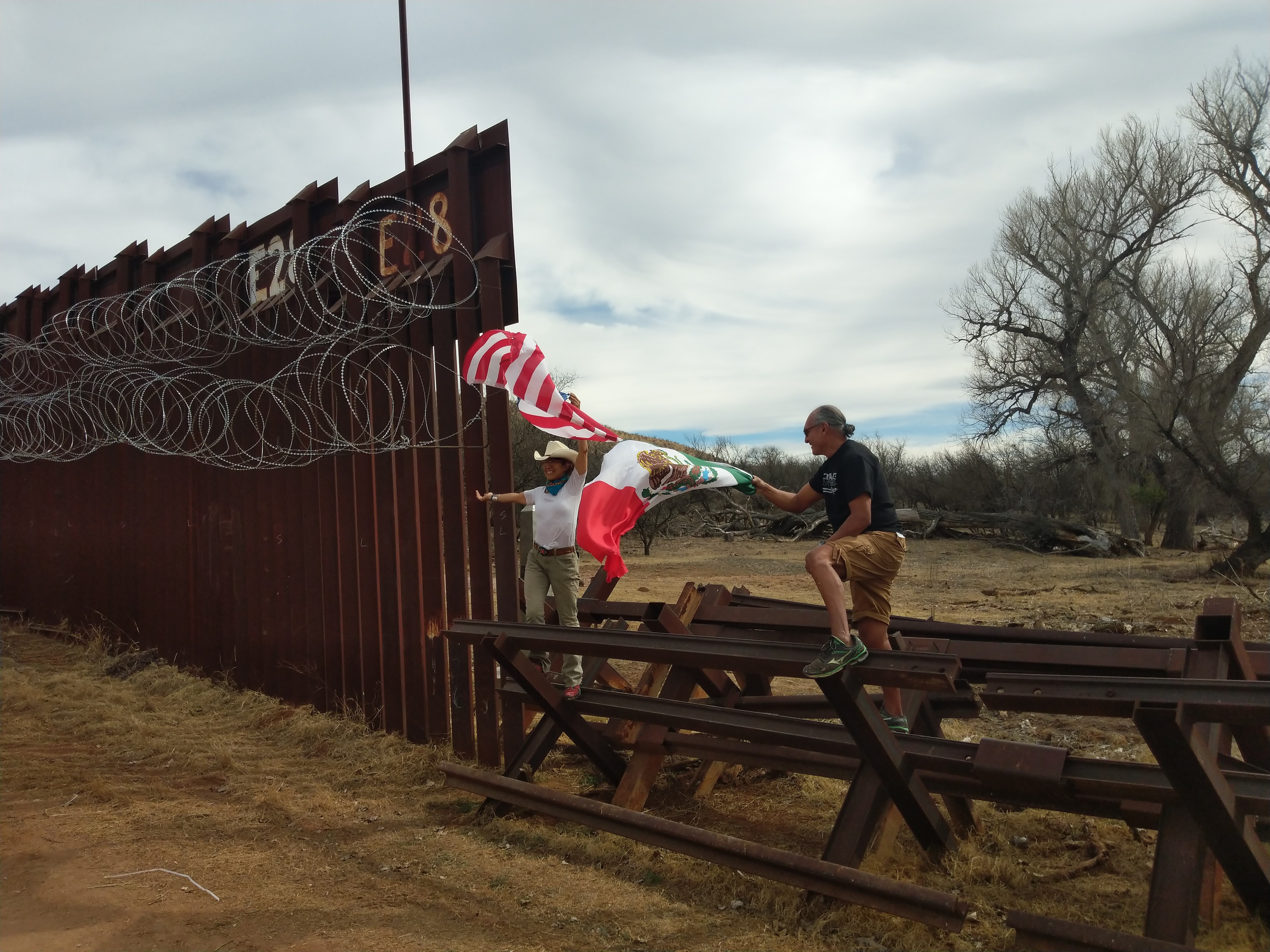 Santa Cruz River — Raechel Running, photographer and activist, and Ricardo Santos Hernández stand on the tank trap barriers at the Santa Cruz River's point of entry into Arizona, the American and Mexican flags flying together in the wind against the rusted steel bollards and coils of razor wire. Two people. Two flags. One border. A defiant act of solidarity at the exact place where two nations — and two peoples who were never meant to be separated — meet. Photography by Raechel Running. by Ricardo Santos Hernández