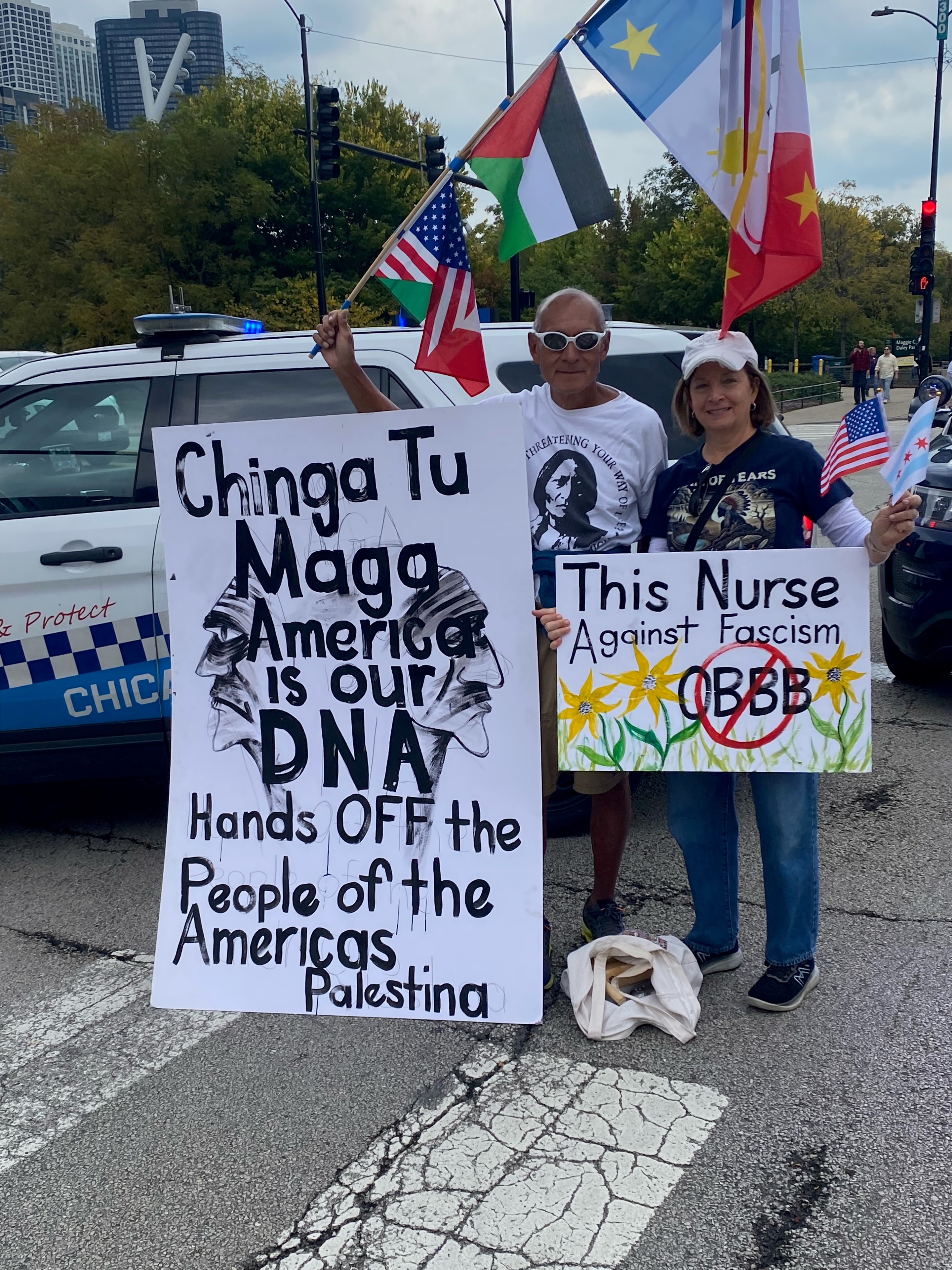 This Nurse Against Fascism — Ricardo Santos Hernández, Chicago, Spring 2026. A photograph from the No Kings March through the streets of downtown Chicago. Ricardo stands alongside a nurse holding her hand-painted sign — "This Nurse Against Fascism" — adorned with sunflowers and a crossed-out OBBB. Ricardo holds his banner: "Chinga Tu MAGA / América is our DNA / Hands OFF the People of the Américas / Palestina." Flags of many nations — Palestinian, Mexican, American, and others — rise behind them. A living document of solidarity: nurses, artists, and people of the Americas marching together against fascism in the streets of Chicago. by Ricardo Santos Hernández