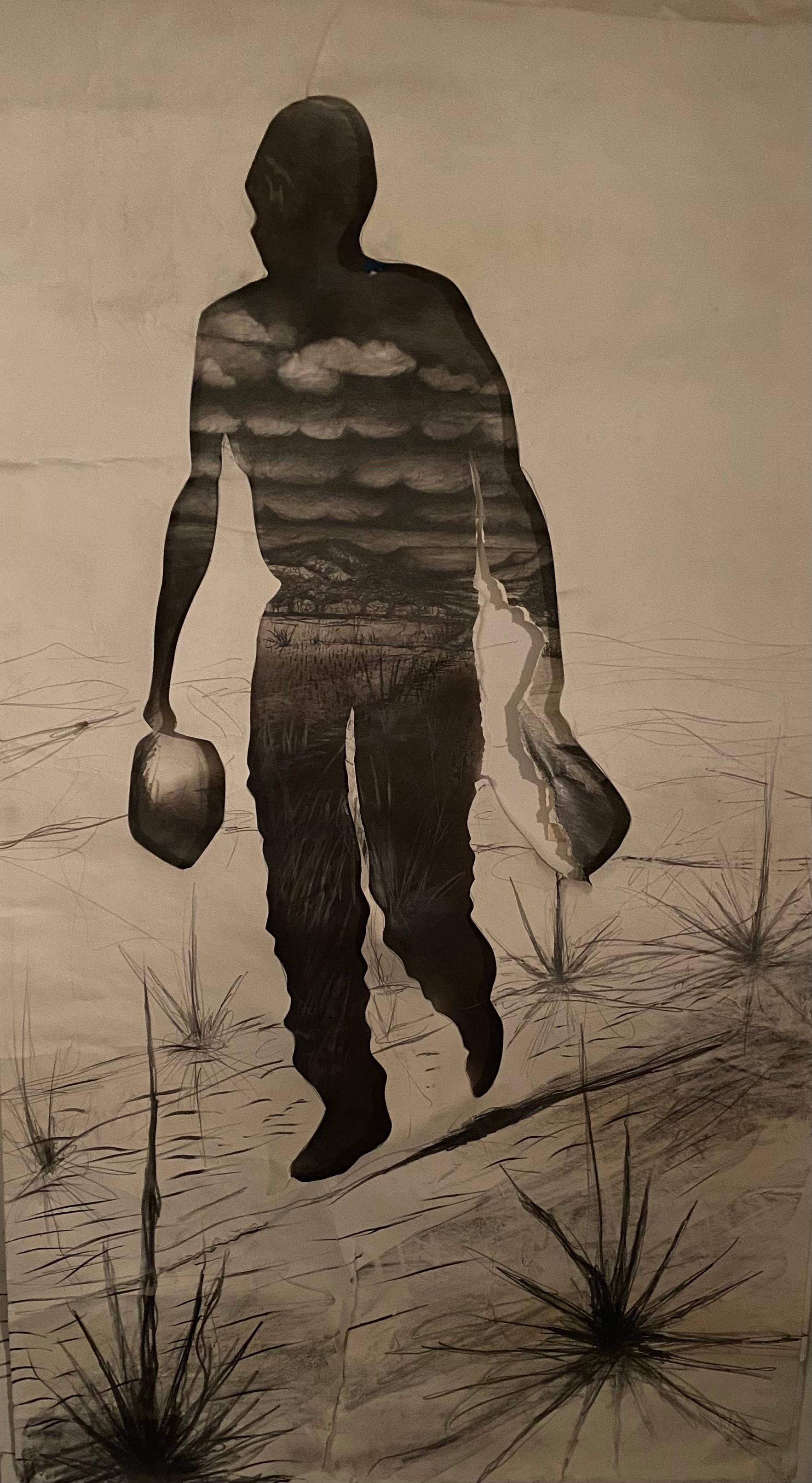 Hombre Cósmico: He Knows the Secret of the Trails — Ricardo Santos Hernández. Large-scale drawing. Graphite sticks, graphite powder on Arches. The silhouette of a man in motion — bag in one hand, hat in the other — fills the sheet from edge to edge. Inside his body, the world he carries: storm clouds layered across his chest, a desert valley stretching through his torso, the horizon line of mountains running through his hips. Around his feet, yucca plants are sketched into the Arches paper as if the earth itself is reaching up to claim him. He is not separate from the land — he is made of it. The trails he knows are written in his body: the sky, the desert floor, the grasses, the long walk. Graphite sticks and powder give the work its weight — the darkness of the silhouette dense and absolute, the landscape within it luminous by contrast. by Ricardo Santos Hernández