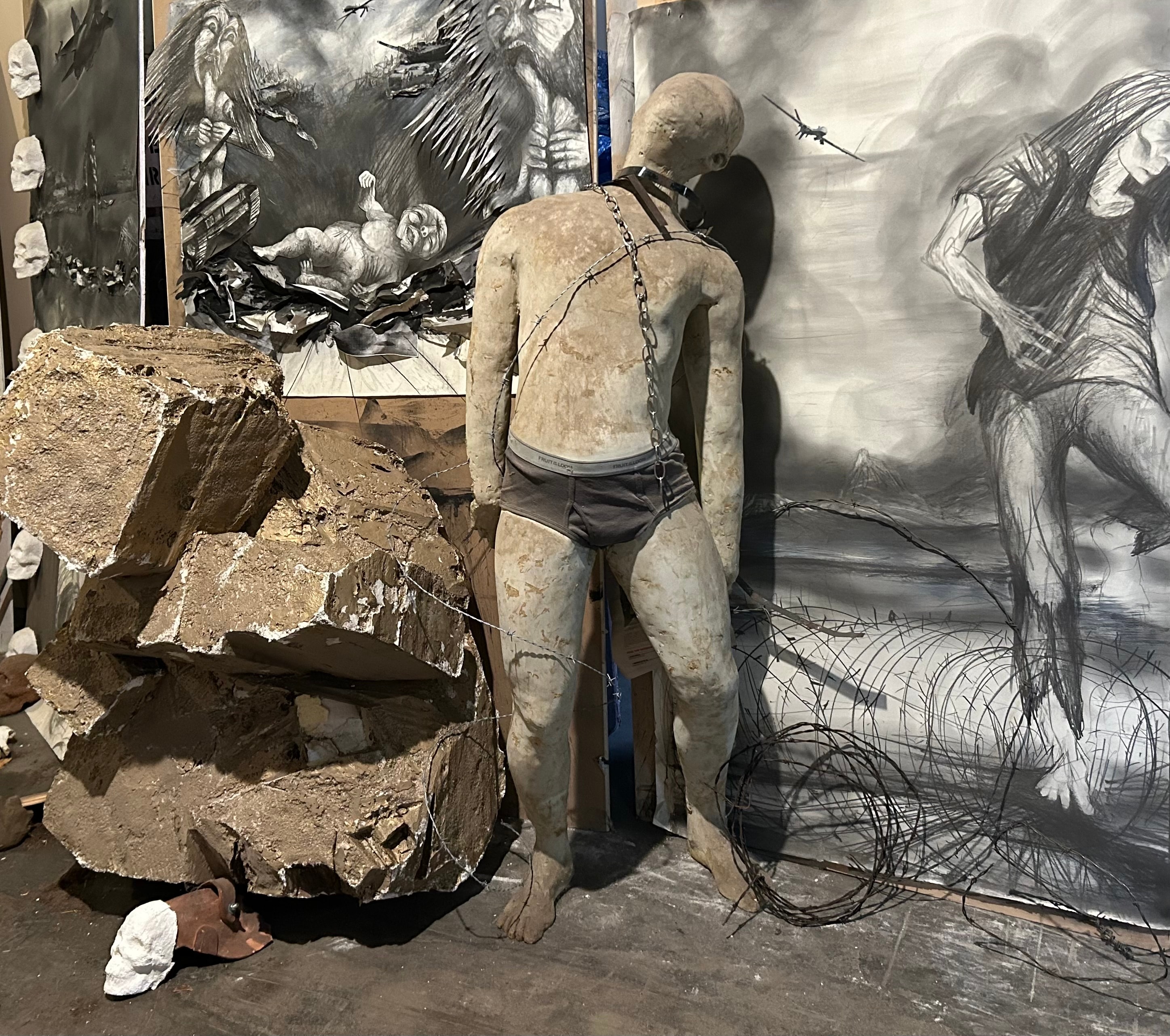 Under the Rubble — Ricardo Santos Hernández, March 27, 2025. A mixed media installation making reference to Christ in the Rubble by Palestinian pastor and theologian Munther Isaac. A figure bound in barbed wire and chains stands amid concrete rubble and large-scale charcoal drawings — bearing witness to the devastation in Gaza and the enduring humanity beneath the destruction. by Ricardo Santos Hernández