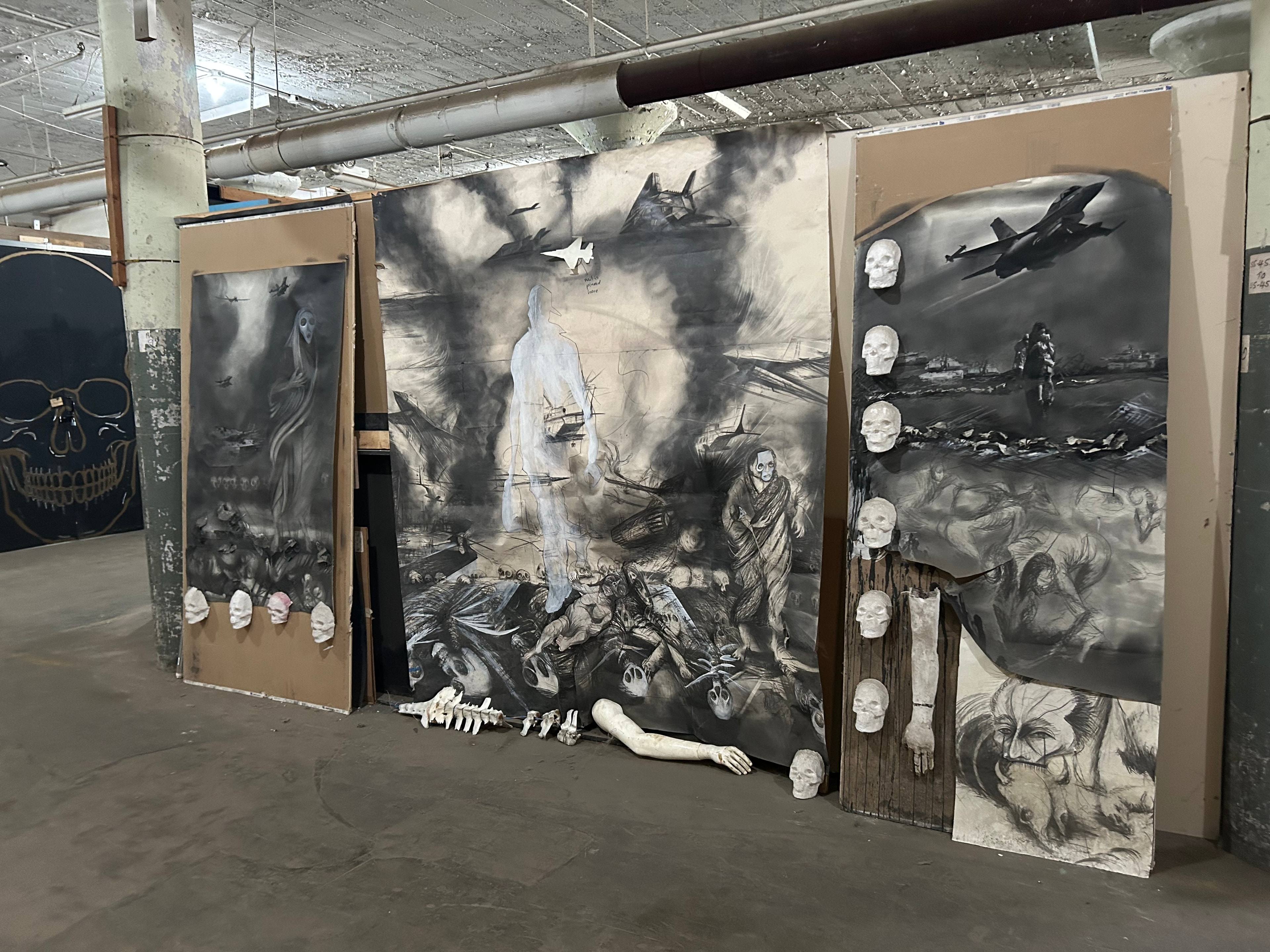 Studio Floor — Large-scale charcoal drawings and mixed-media installation panels in progress on the studio floor. These monumental works — featuring skulls, ghostly figures, and fighter jets — reflect Ricardo's ongoing engagement with war, memory, and resistance.